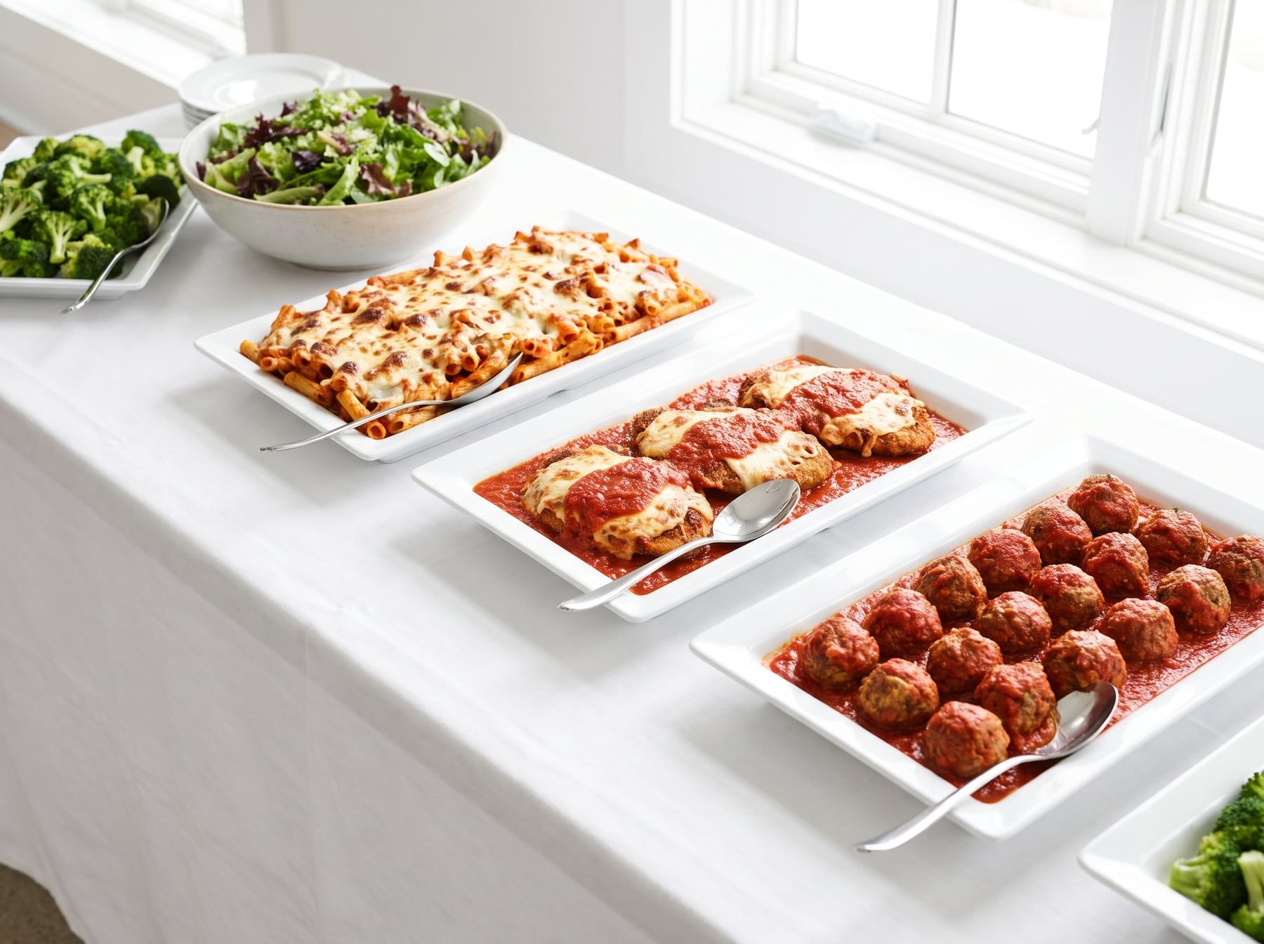 Catering dishes of baked ziti, chicken parmigiana and meatballs on a long white table