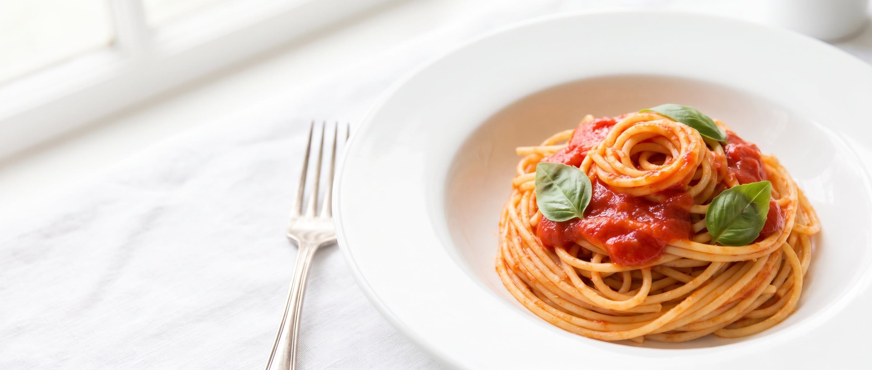 Spaghetti in tomato sauce with fresh basil in a white bowl, on white linen by a window